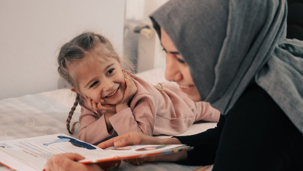Woman in a grey headscarf reading to her daughter on a bed who is looking at her smiling