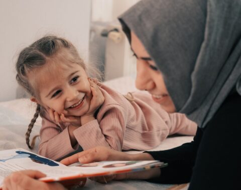 Woman in a grey headscarf reading to her daughter on a bed who is looking at her smiling