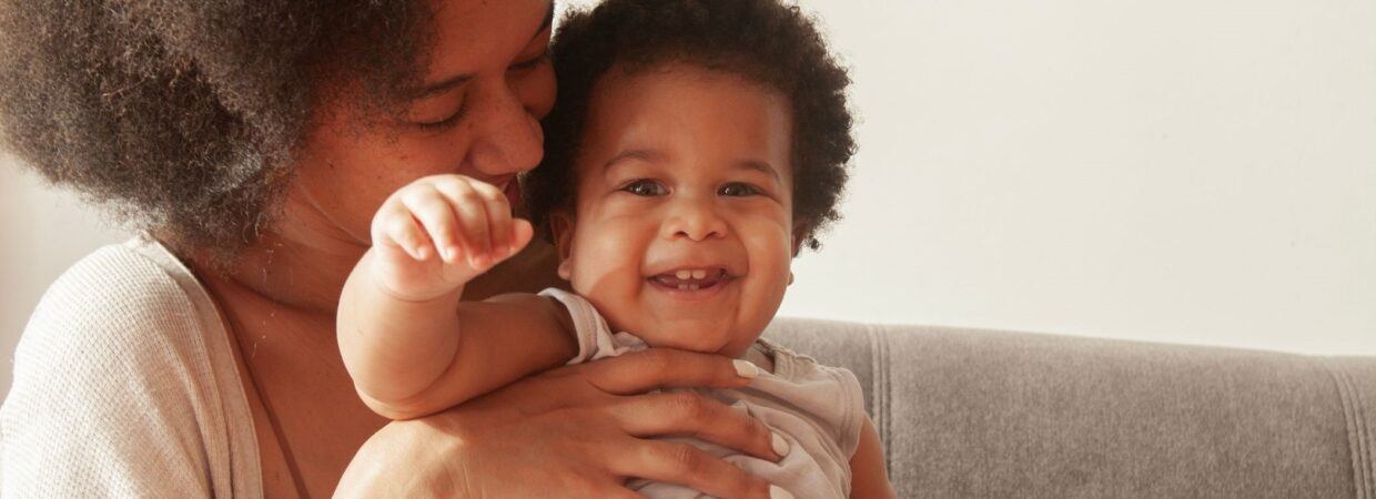 A mother with black afro hair wearing a beige top holding a baby and laughing, sat on a grey sofa