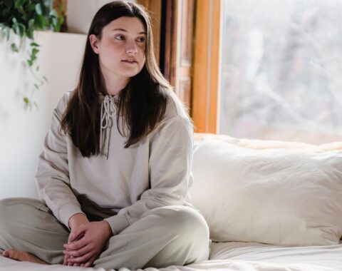 Young woman with long brown hair sat cross-legged on a bed in her own room