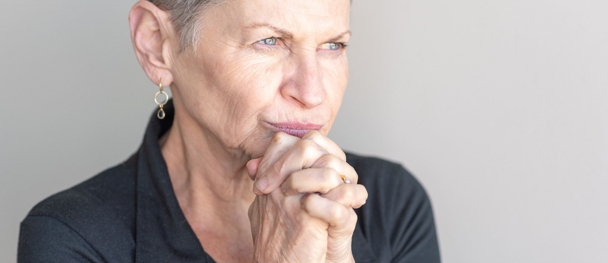 An older woman with hands helf together under her chin, staring into the distance