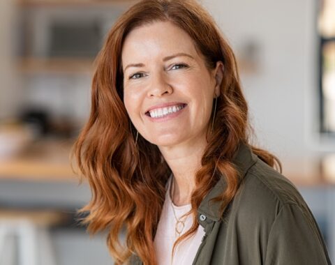 A middle aged woman with long red wavy hair wearing a khaki blouse over a white t shirt in a kitchen smiling