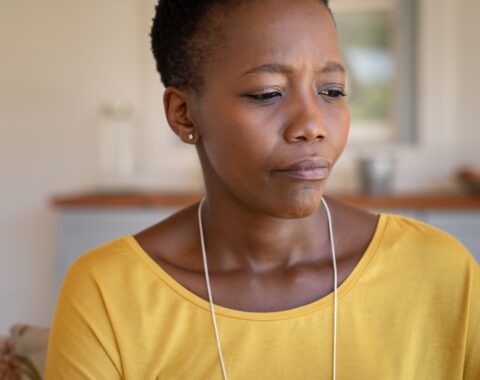 An older woman in yellow top and cropped hair looking down with a thoughtful expression on her face