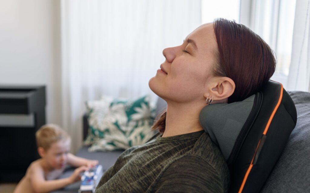 Young woman sat on a sofa leaning head back and smiling, with son in the background