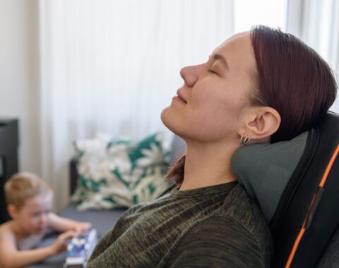 Young woman sat on a sofa leaning head back and smiling, with son in the background