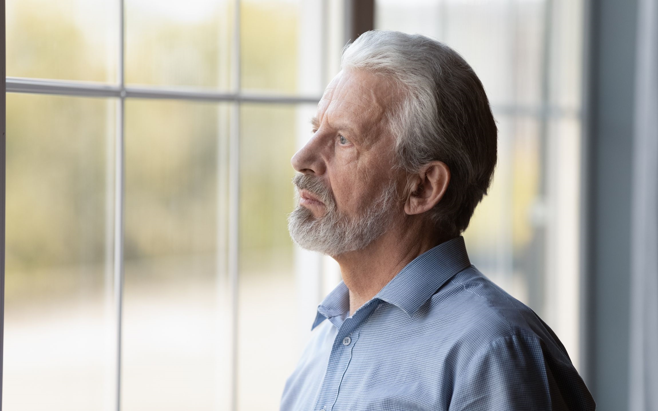Older man with white hair looking out of a window with a sad expression on his face