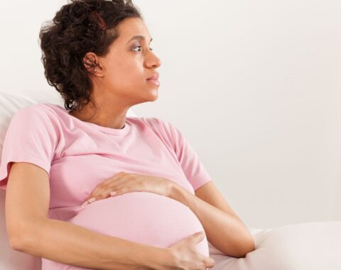 A pregnant woman with curly hair wearing a pink t shirt, holding her pregnant belly