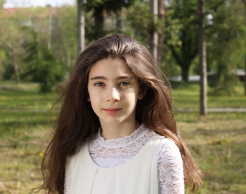 Youn girl with long dark hair wearing a white top, standing outdoors looking to camera