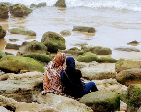 Two women sat on beach rocks comforting each other looking out to sea