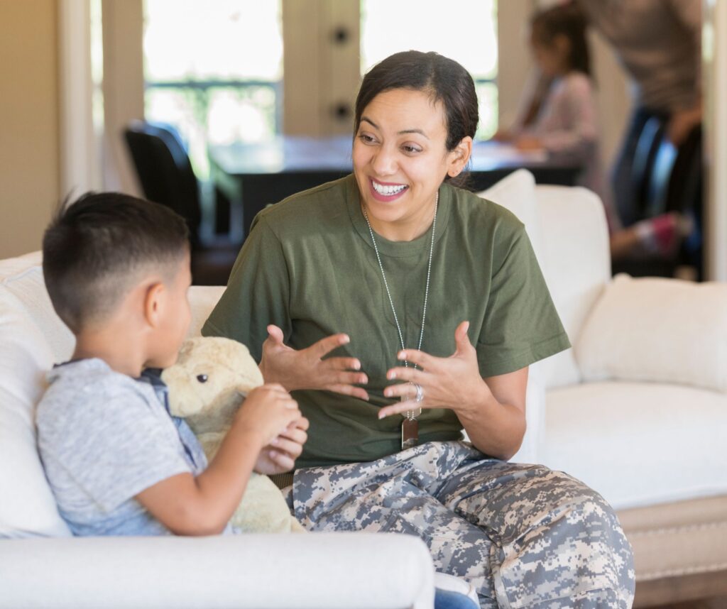 Young mother sat on a sofa with her young son who is cuddling a teddy, explaining something and smiling.