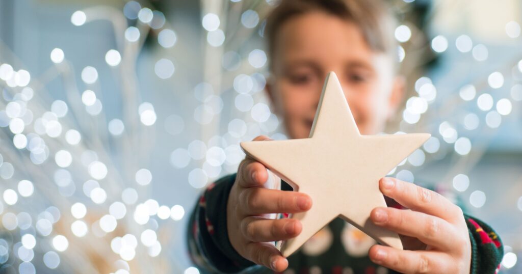 A young boy stands in front of Christmas lights with his arms outstretched, holding a Christmas star in front of him. He wears a green Christmas jumper, has blonde hair and is smiling.