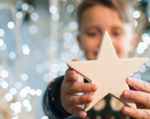 Young boy standing in front of Christmas lights holding a star out in front of him smiling. He wears a Christmas jumper and has short blonde hair.