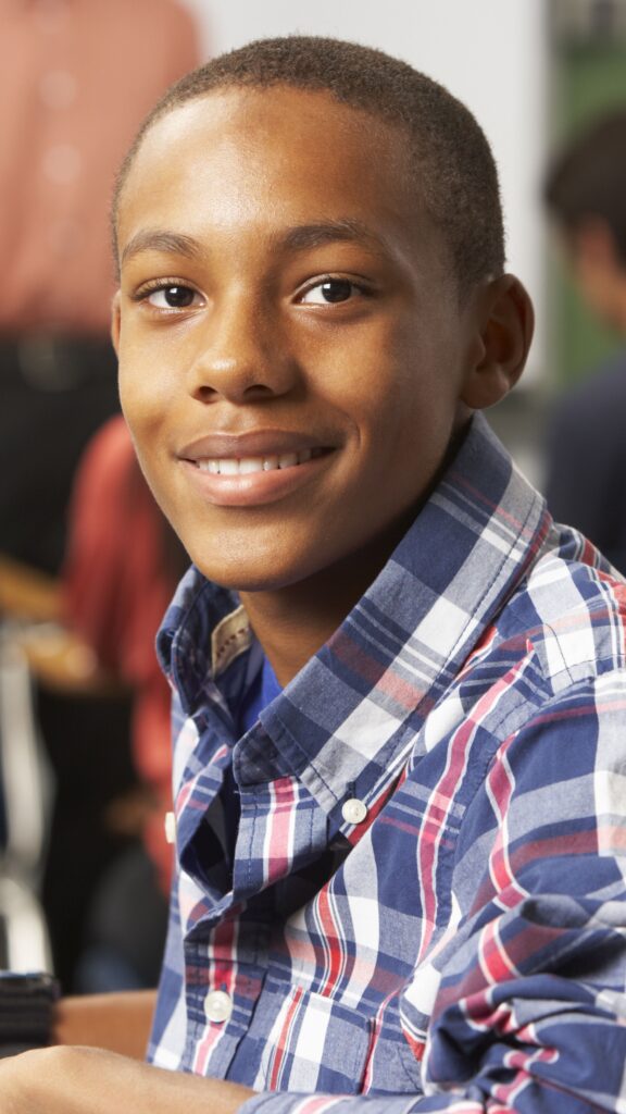A black Boy in a classroom looking forward and smiling. He wears a blue, red and white checked long-sleeve shirt